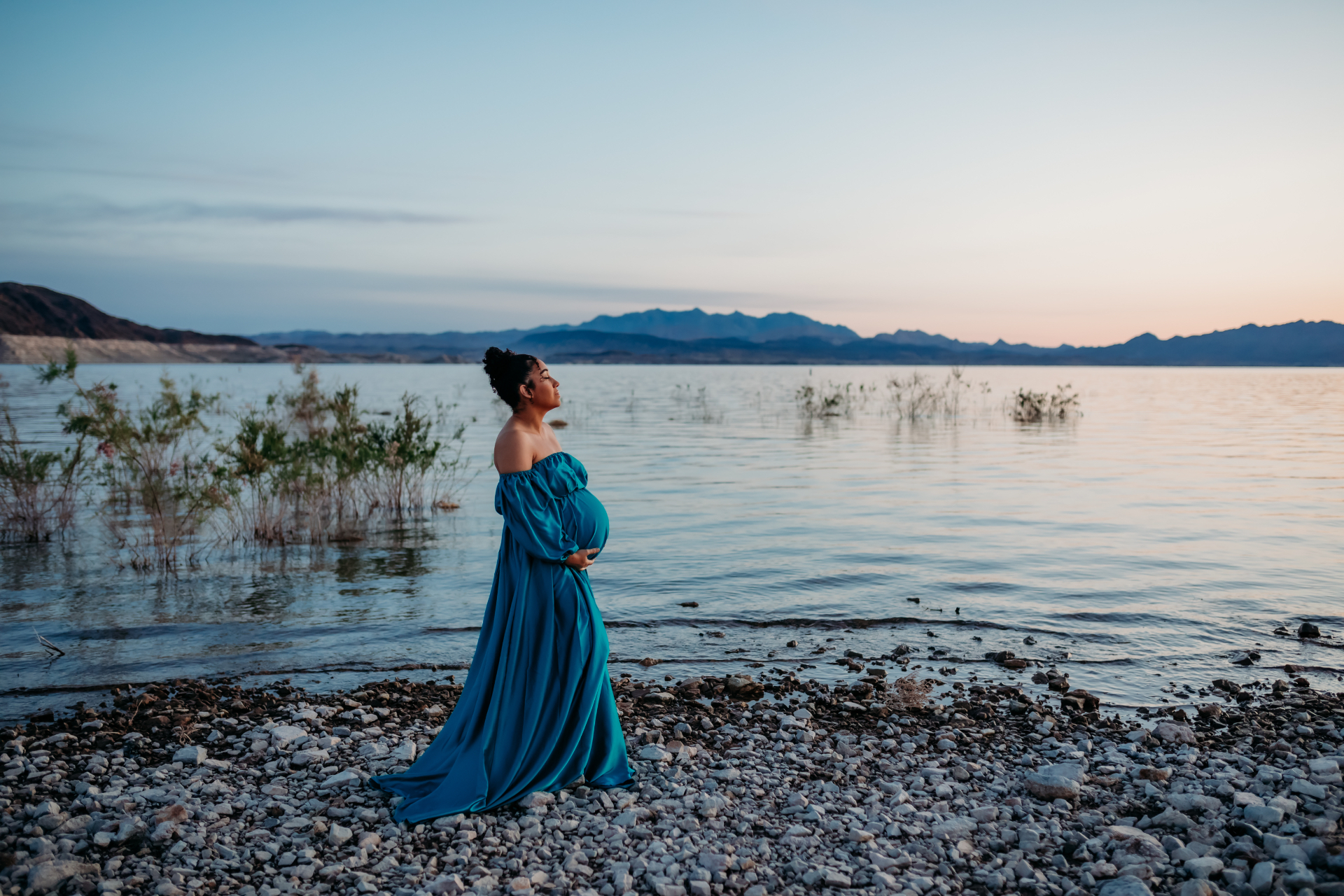A pregnant woman in a flowing teal dress standing by a calm lake at sunset
